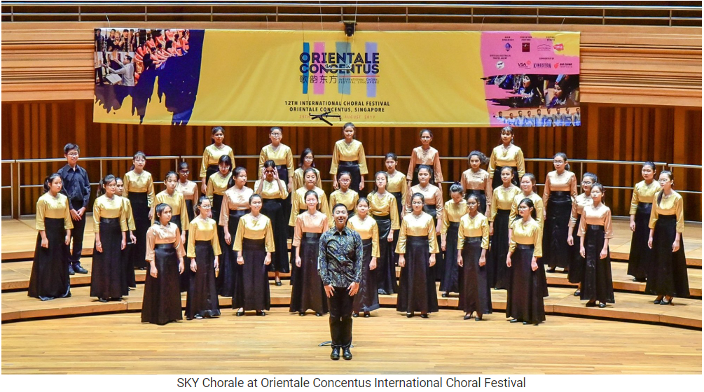 A choir in gold tops and black skirts stands on a wooden stage at the Orientale Concentus International Choral Festival.